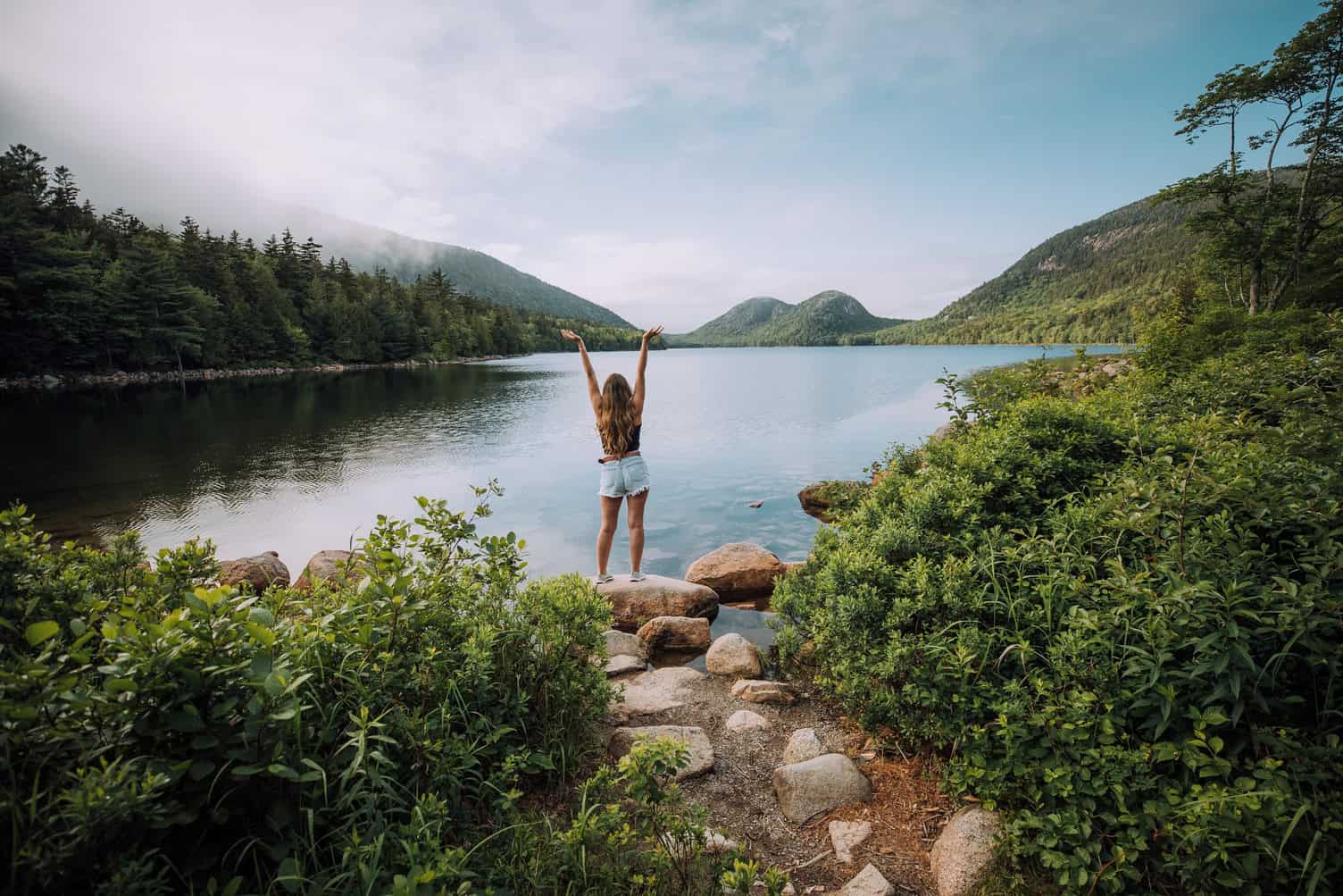 Woman enjoying nature by a lake surrounded by mountains and greenery.