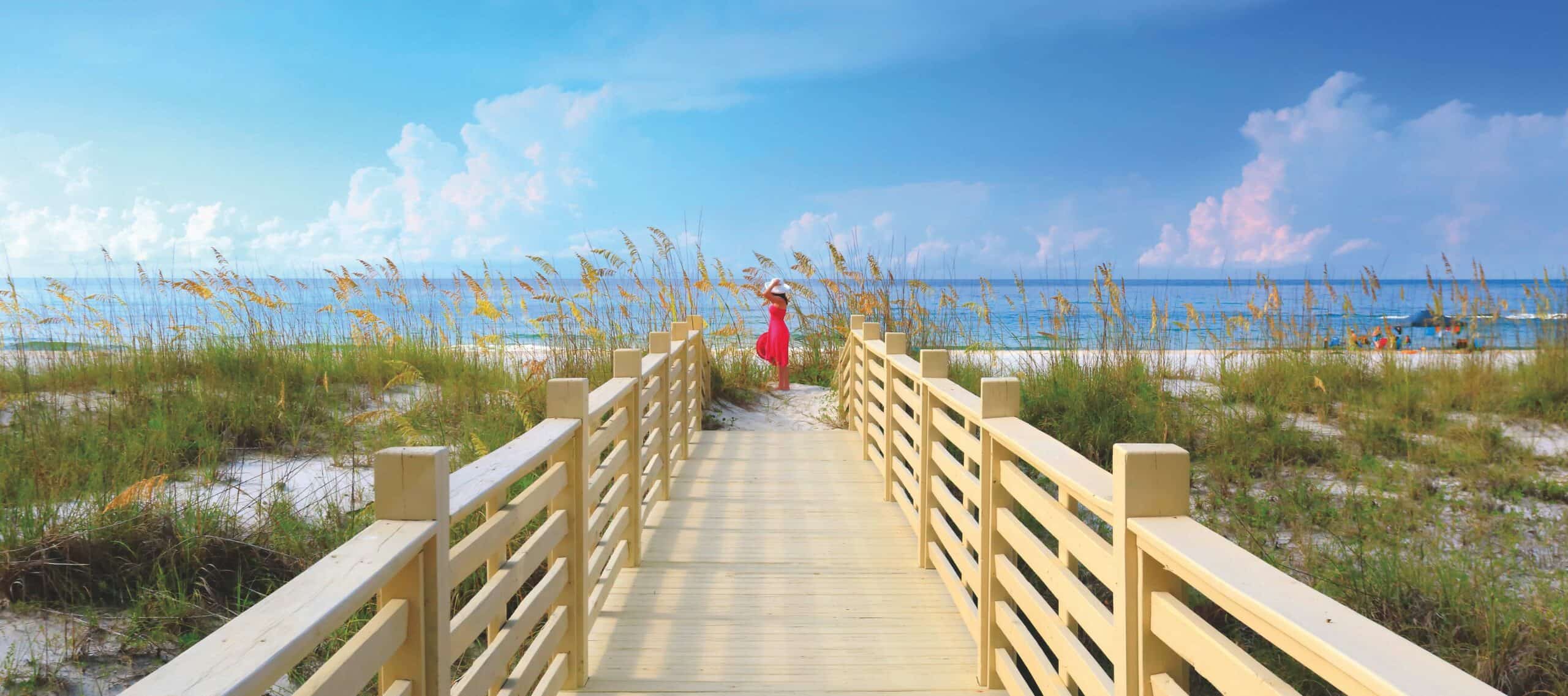 Wooden pathway leading to a sandy beach and blue ocean under a partly cloudy sky.