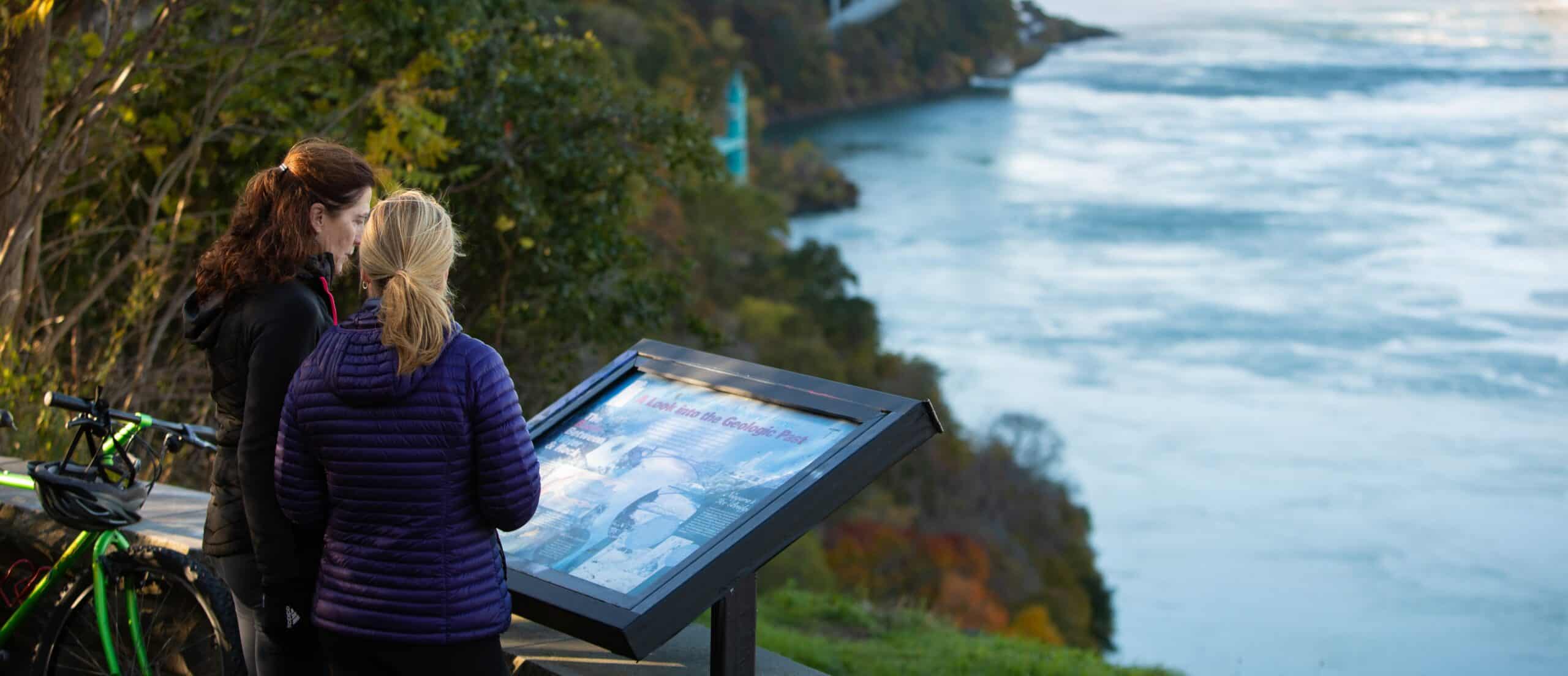 Two women looking at an outdoor map overlooking a river and forest.
