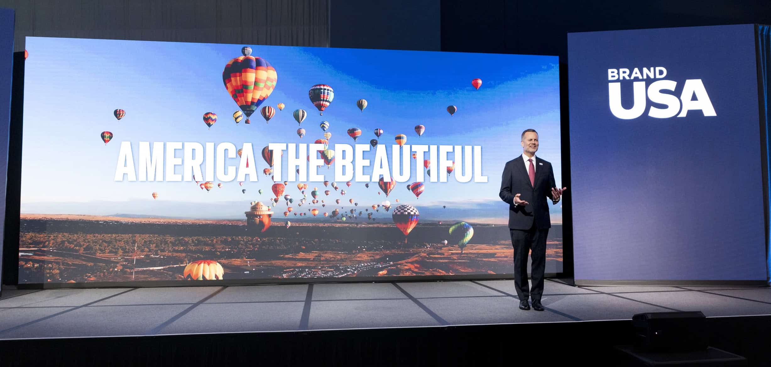 Speaker presenting at Brand USA event with hot air balloons in background.