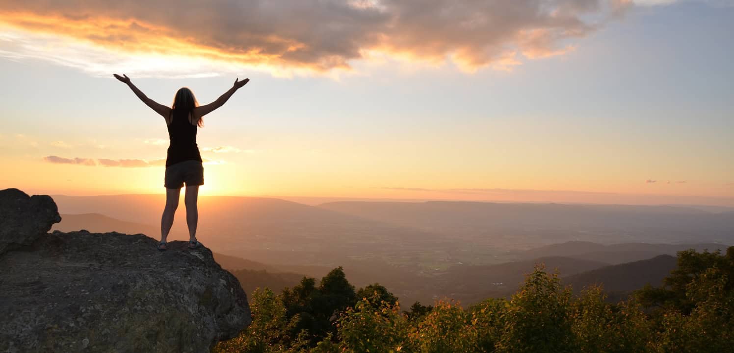 Woman standing on a rock with arms raised at sunset in nature.