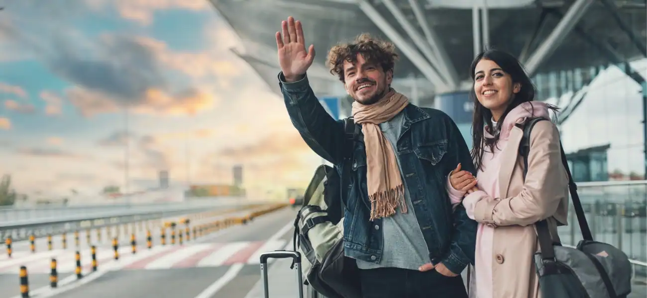 Smiling travelers waving at airport terminal with luggage at sunset.