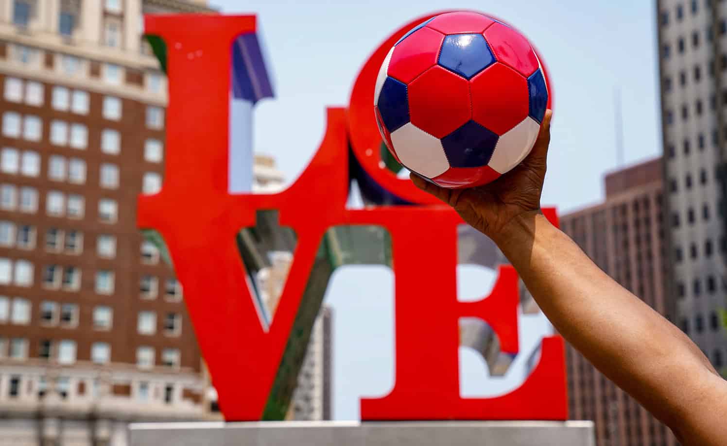 Person holding a soccer ball in front of the LOVE sign in an urban setting.